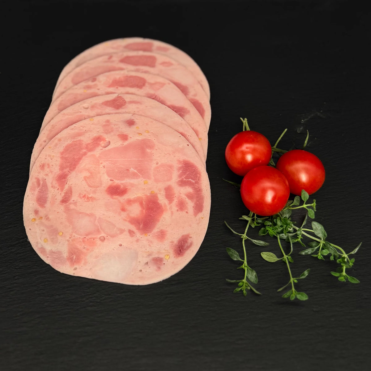 Geschnittener Bierschinken gefächert, auf einer schwarzen Schieferplatte mit Dekoration aus Tomaten und Kräutern.