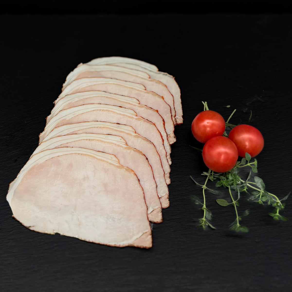 Geschnittener Bratenaufschnitt, gefächert, auf einer schwarzen Schieferplatte mit Dekoration aus Tomaten und Kräutern.