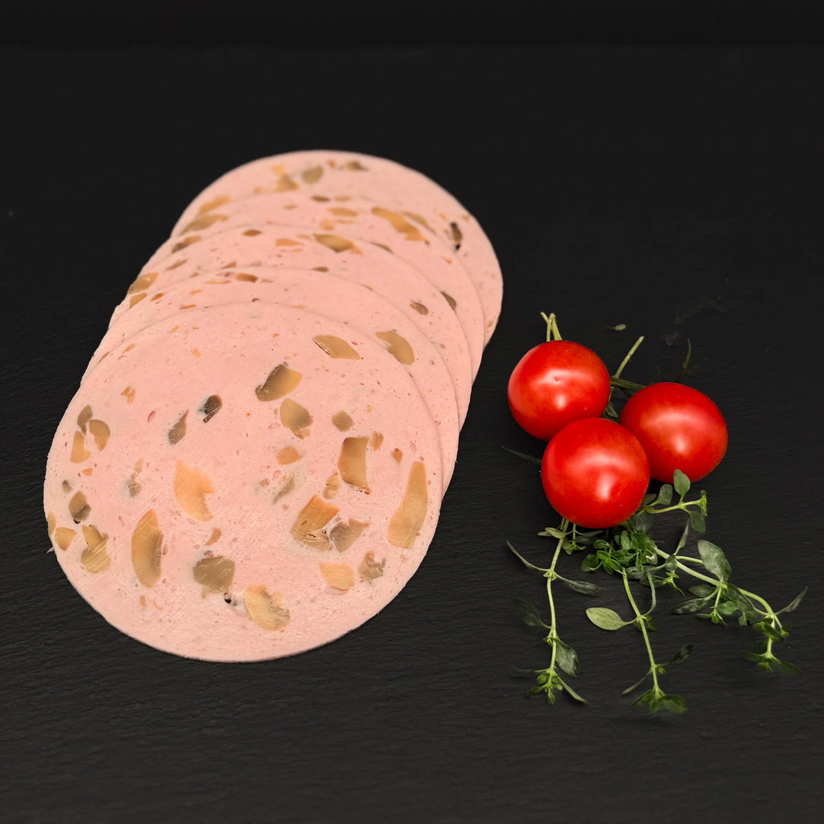 Geschnittene Champignon Mortadella, gefächert, auf einer schwarzen Schieferplatte mit Dekoration aus Tomaten und Kräutern.