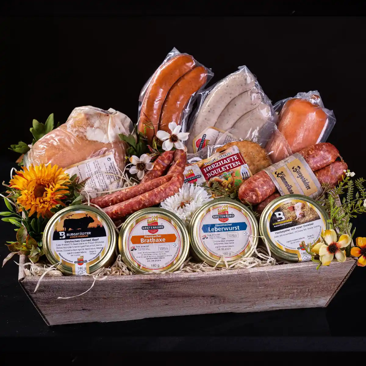 Ein rechteckiger Holzkorb gefüllt mit vier Gläsern, Pfefferbeissern, zwei Kümmelknackern, verschiedneen anderen Wurstsorten in Vakuumfolie wie Bouletten, Rauchwurst, Roster/Bratwurst, Jagdwurst und der Grillhaxe.