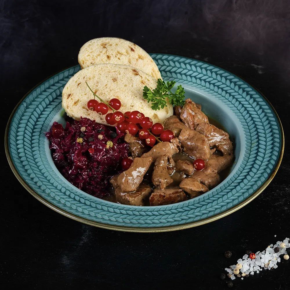 Blauer Teller mit Hirschgulasch, Blaukraut und zwei Klößen vor dunklem Hintergrund.