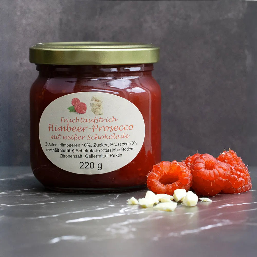 Roter Fruchtaufstrich im Glas neben Himbeeren und weißen Schokoladenstückchen vor dunkelgrauem Hintergrund.