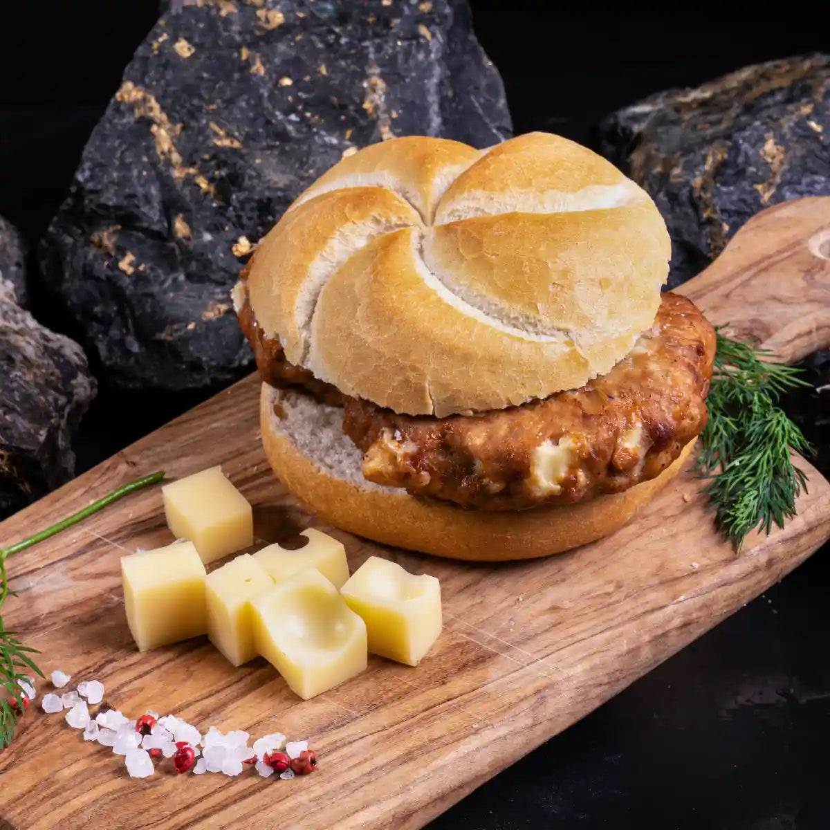 Ein helles Brötchen mit gebratener Boulette auf einem Holzbrett daneben einige Käsewürfel als Dekoration