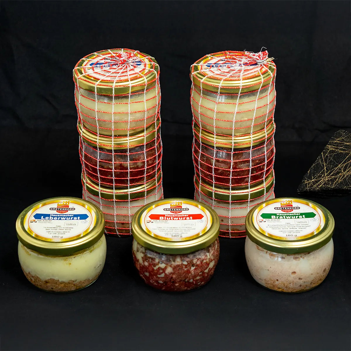 Drei Wurstgläser vor zwei Türmen aus jeweils drei Wurstgläsern in Netzen vor dunklem Hintergrund.