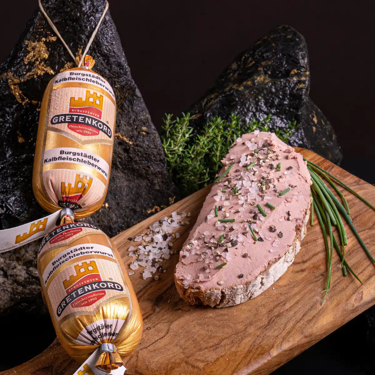 Kalbfleischleberwurst im Darm und daneben auf einem dunklen Holzbrett ein Brot, bestrichen mit Kalbfleischleberwurst und mit Schnittlauch und garniert.