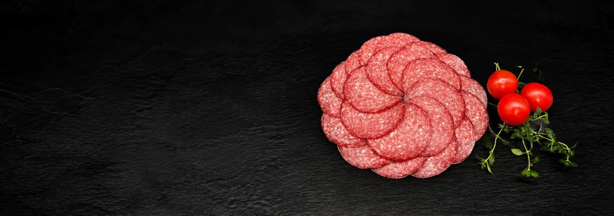 Geschnittene Hausmacher Salami, als Kreis gelegt, auf einer schwarzen Schieferplatte mit Dekoration aus Tomaten und Kräutern.