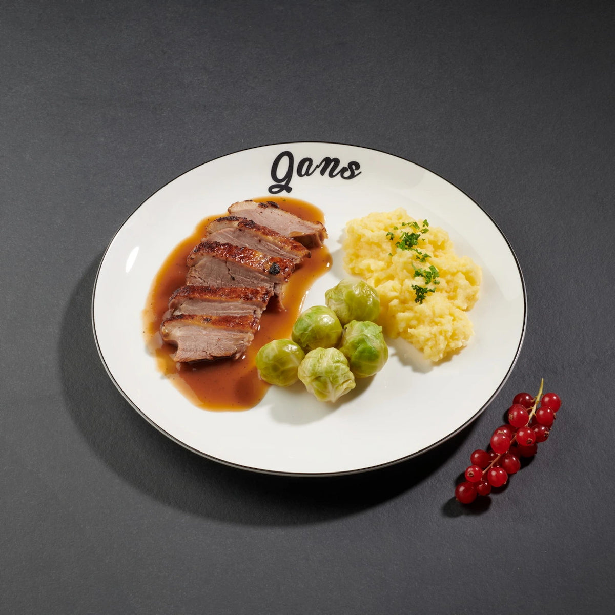 Scheiben von Gänsebrust mit Soße, Rosenkohl und Kartoffelstampf mit Petersilie angerichtet auf einem weißen Teller. An der Seite befinden sich rote Johannisbeeren als Deko.