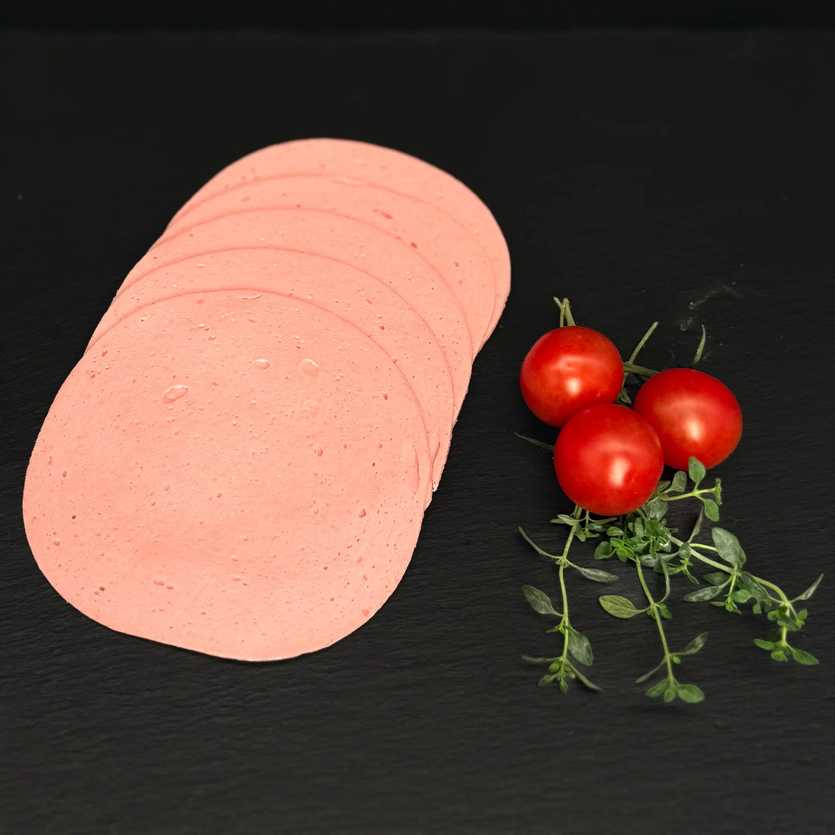 Geschnittener Leberkäse, gefächert, auf einer schwarzen Schieferplatte mit Dekoration aus Tomaten und Kräutern.