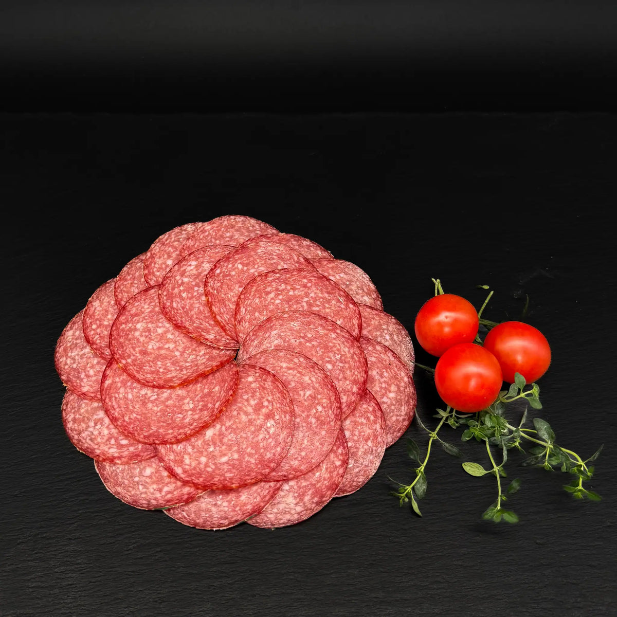 Geschnittene Hausmacher Salami, als Kreis gelegt, auf einer schwarzen Schieferplatte mit Dekoration aus Tomaten und Kräutern.