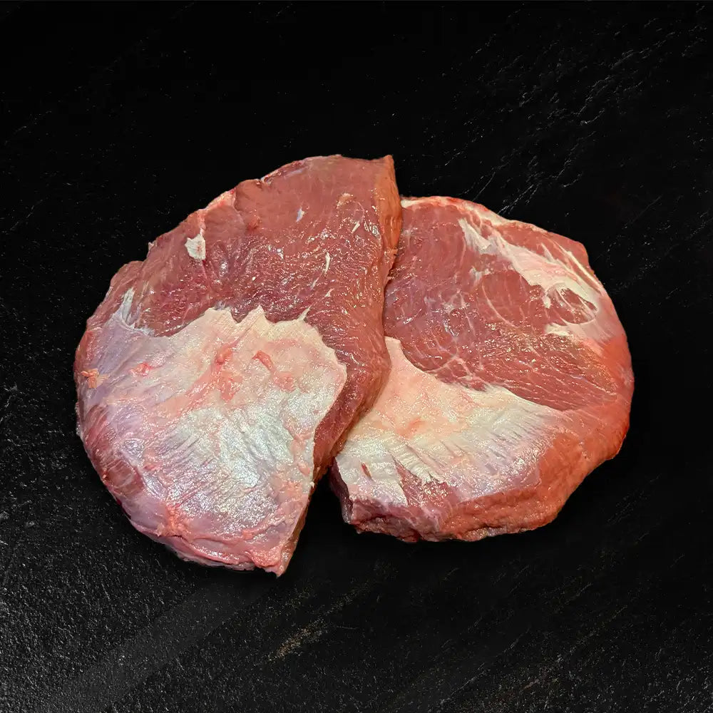 Zwei Kalbsbäckchen liegen nebeneinander auf dunklem Hintergrund.