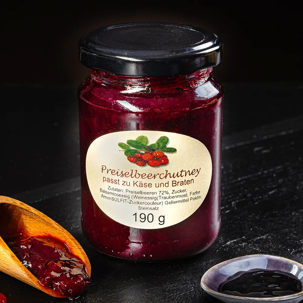 Dunkelrotes Preiselbeerchutney im Glas neben kleinem Holzlöffel und Schälchen mit Sauce vor dunklem Hintergrund.