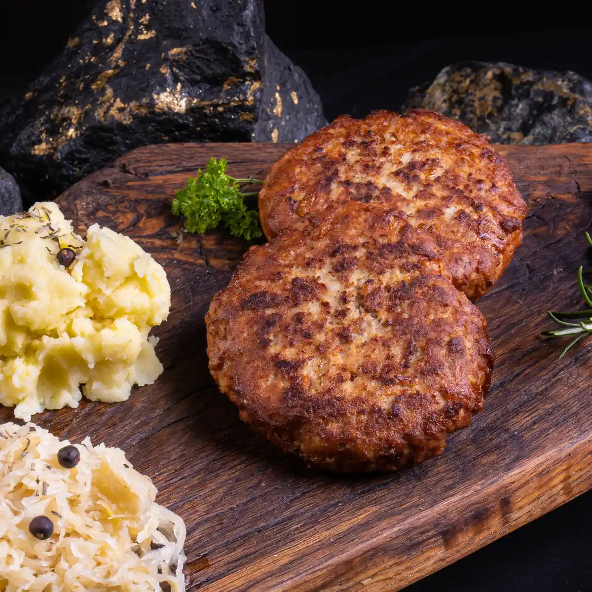 Zwei gebratene Bouletten auf einem dunklen Holzbrett daneben mit Kartoffelsalat und Sauerkraut garniert