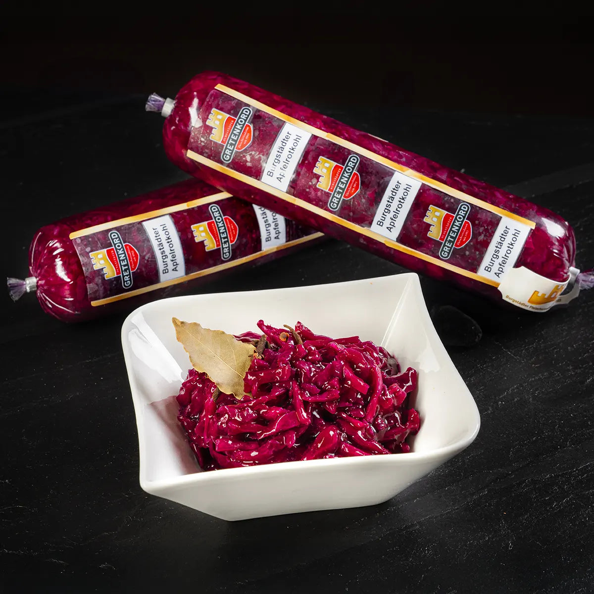Burgstädter Apfelrotkohl in weißem Schälchen mit zwei Packungen Apfelrotkohl vor dunklem Hintergrund.