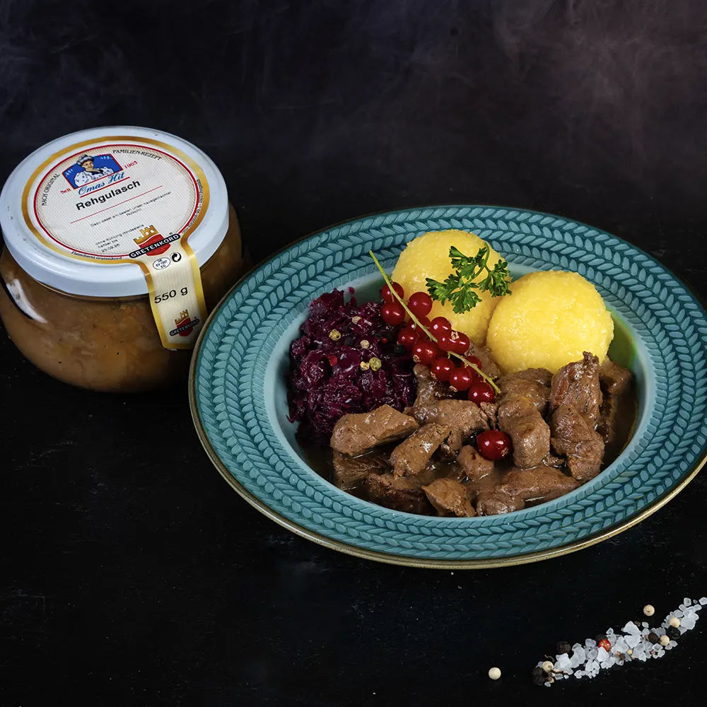 Blauer Teller mit reggulasch , Rotkraut und zwei Klößen neben Glas mit Rehgulasch.