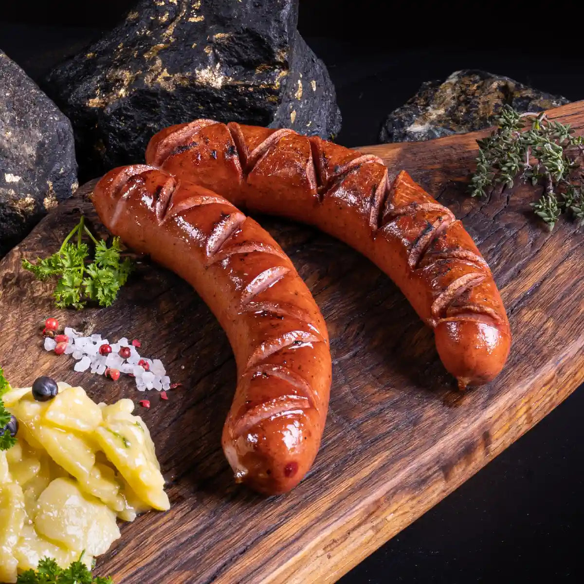 Zwei gegrillte und eingeschnittene Rauchwürste auf einem Holzbrett mit Kartoffelsalat daneben