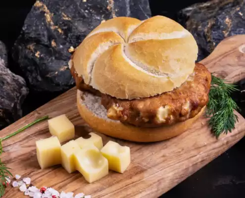 Ein helles Brötchen mit gebratener Boulette auf einem Holzbrett daneben einige Käsewürfel als Dekoration